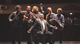 Eine Gruppe von sechs Musikern auf einer Bühne. Sie spielen verschiedene Instrumente und haben Spaß beim Musizieren.