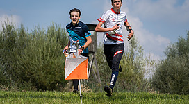 HSV Ried Orientierungslauf in Ried im Innkreis: Zwei junge Läufer in Sportausrüstung laufen durch eine grüne Umgebung mit Bäumen und einem blauen Himmel im Hintergrund. Der eine Läufer hält eine orangefarbene Fahne, die auf eine Veranstaltung oder einen Wettkampf hinweist.