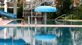 Erlebnis-Freibad Enns in Enns: Eine Außenaufnahme eines Swimmingpools mit blauem Wasser und einem hohen Springturm. Sonnenschirme und Liegestühle sind rund um den Pool aufgestellt. Im Hintergrund sind mehrgeschossige Gebäude mit Balkonen zu sehen.