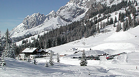 Eine verschneite Berglandschaft mit hohen Bergen und einem klaren blauen Himmel. Im Vordergrund steht ein kleines Gebäude, umgeben von tiefem Schnee und Nadelbäumen.