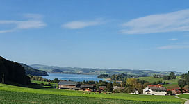 Aussichtsplatz Hochgrub in Lochen: Eine idyllische ländliche Landschaft mit grünen Wiesen, Bäumen und Häusern. In der Ferne ist ein See oder Fluss zu sehen, der von bewaldeten Hügeln und Bergen umgeben ist. Der Himmel ist wolkenlos und blau. Die Szenerie vermittelt eine friedliche und ruhige Atmosphäre.