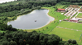 Sportbuffet in Steyregg: Eine weite, grüne Landschaft, in der sich ein großer See befindet. Am Ufer des Sees befinden sich mehrere Gebäude, die offensichtlich einer touristischen Einrichtung zugehörig sind. Die Umgebung ist von Wäldern und Wiesen geprägt, was auf eine ländliche, naturnahe Umgebung hindeutet.