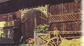 Bauern- u. Heimatmuseum in Haigermoos: Eine ländliche Szenerie mit einem alten Holzgebäude, das den Schriftzug "Kultur & Brauchstum" trägt. Vor dem Gebäude befinden sich Ballen Heu und ein traditionell anmutendes Fuhrwerk. Ein Paar sitzt entspannt auf den Heuballen und scheint den Anblick zu genießen.