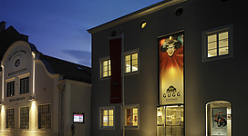 Gugg Kulturhaus in Braunau am Inn: Ein mehrstöckiges Gebäude mit beleuchteten Fenstern und einer Eingangstür. An der Fassade befindet sich ein großes Schild mit dem Namen "Gugg". Es scheint sich um ein kulturelles Zentrum oder eine ähnliche Einrichtung zu handeln.
