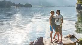 Wolfgangsee in St. Wolfgang im Salzkammergut: Das Bild zeigt ein junges Paar, das auf einem Holzsteg an einem See stehend, die malerische Landschaft mit Bergen und einem Ausflugsschiff im Hintergrund betrachtet. Die Umgebung vermittelt eine friedliche und entspannende Atmosphäre.