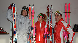 Drei Skifahrer posieren mit bunten Skiern in einem Raum. Sie tragen Skikleidung und haben Skibrillen auf.