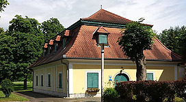 Historisches Schützenhaus in Enns: Ein charmantes Gebäude mit tradtioneller Architektur, umgeben von üppiger Vegetation. Das zweigeschossige Haus mit rotem Ziegeldach und gelbem Putz steht in einer idyllischen grünen Umgebung mit Bäumen und Sträuchern.