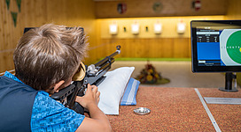 Ein Junge zielt mit einem Luftgewehr auf eine Zielscheibe in einer Schießhalle. Im Hintergrund ist ein Bildschirm mit den Schießdaten sichtbar.