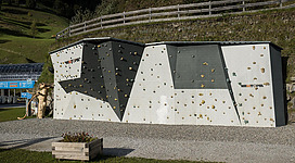 Eine Kletterwand mit verschiedenen Griffen und Strukturen. Im Hintergrund sind grüne Hügel und eine Holzplanke zu sehen.