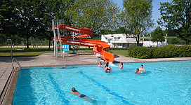 Freibad Andorf in Andorf: Eine Wasserrutsche und ein Schwimmbecken in einer parkähnlichen Umgebung. Es sind mehrere Menschen unterschiedlichen Alters beim Schwimmen und Rutschen zu sehen. Die Anlage erscheint gepflegt und einladend.