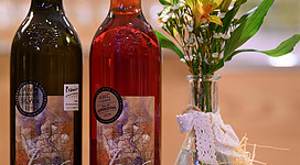 Zwei Flaschen Wein, eine weiße und eine rosé, stehen auf einem Tisch. Neben den Flaschen befindet sich eine kleine Vase mit frischen Blumen.