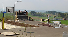 Ein Skatepark mit verschiedenen Rampen und einer Basketball-Arena. Mehrere Jugendliche fahren Skateboard und verbringen Zeit im Freien.