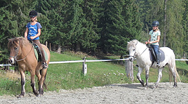 Zwei Mädchen reiten auf Ponys in einer grünen Umgebung. Im Hintergrund sind Bäume zu sehen.