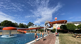Freibad Mauthausen in Mauthausen: Offensichtlich ein Erholungsort mit einem Schwimmbad, verschiedenen Rutschen und einer lebendigen Umgebung mit Besuchern. Es gibt üppigen Grünbewuchs, einen blauen Himmel mit Wolken und eine attraktive Architektur des Gebäudes.