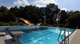 Erlebnisbad Freibad Zell am Pettenfirst in Zell am Pettenfirst: Ein Außenbereich mit einem großen Schwimmbecken, umgeben von Bäumen und blauem Himmel. Es gibt mehrere Plattformen und Rutschbahnen, die in das Becken führen. Die Wasseroberfläche ist klar und einladend.