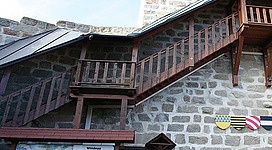 Burgruine Windegg in Schwertberg: Ein mehrstöckiges Gebäude aus grauem Stein mit hölzernen Balkonen und Veranden. Das Dach hat eine traditionelle Bauweise mit vorfragenden Dachspitzen. An den Wänden sind verschiedene Wappen und Schilder angebracht, die auf eine geschichtliche Bedeutung des Ortes hinweisen.