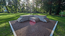 Ein moderner Sitzbereich aus Beton in einem grünen Park. Die Sonne scheint durch die Bäume und schafft eine angenehme Atmosphäre.
