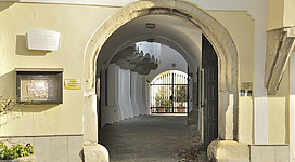 Landesfürstliche Burg in Enns: Ein gewölbter Durchgang führt zu einer Terrasse mit Metallgeländer. Das Gebäude hat eine alte, verwitterte Fassade und traditionelle Architektur. Im Hintergrund sieht man eine idyllische Umgebung mit üppiger Vegetation. Der Ort vermittelt einen historischen und gemütlichen Charakter.