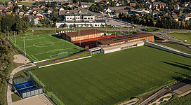 Ein modernes Sportgelände mit Fußballfeldern und Tennisplätzen. Im Hintergrund sind Wohngebäude und eine natürliche Umgebung sichtbar.