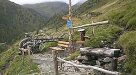 Ein Wanderweg in den Bergen mit Holzschildern und Bänken. Die Umgebung ist grün und hügelig, mit bewölktem Himmel.