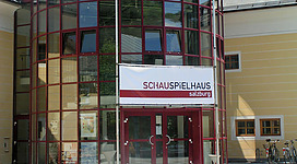 <p>Ein modernes Gebäude mit rot umrandetem Glas und einer großen Eingangstür. Über dem Eingang hängt ein Schild mit dem Schriftzug "SCHAUSPIELHAUS Salzburg".</p>
