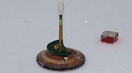 Ein Holzspielstein steht auf einer weißen, schneebedeckten Fläche. Daneben liegt ein roter Würfel.