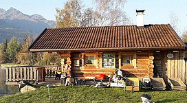 Ein gemütliches Holzhaus am Wasser mit einer schönen Aussicht auf die Berge. Die Umgebung ist grün und einladend, ideal für einen entspannten Aufenthalt.