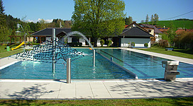 Freibad Oberkappel in Oberkappel: Ein Außenpool-Komplex umgeben von grüner Vegetation. Im Zentrum befindet sich ein großer, rechteckiger Pool mit Sprungbrett und Wasserrutsche. Mehrere kleine Gebäude mit Dächern in traditioneller Bauweise und hellen Fassaden säumen den Pool. Bäume mit dichter Belaubung rahmen die Szene ein.