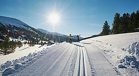 Ein sonniger Wintertag in den Bergen mit zwei Personen auf einer präparierten Langlaufloipe. Die Landschaft ist von schneebedeckten Bergen und Tannenbäumen umgeben.