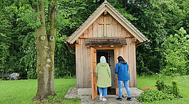 Siebenschläferkapelle in Munderfing: Eine hölzerne Kapelle inmitten üppiger Bäume und Vegetation. Zwei Personen stehen davor und scheinen sie zu betreten.