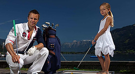 Ein Mann und ein Mädchen spielen Minigolf am Wasser. Im Hintergrund sind Berge und ein klarer Himmel zu sehen.