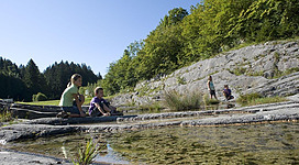 Kinder spielen am Ufer eines Gewässers in der Natur. Im Hintergrund sind grüne Bäume und felsige Wände zu sehen.
