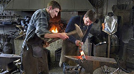 Zwei Schmiede arbeiten in einer traditionellen Schmiede. Einer schlägt mit einem Hammer auf ein glühendes Stück Metall, während der andere es formt.