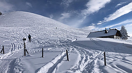 Eine verschneite Landschaft mit einem kleinen Haus und mehreren Fußspuren im Schnee. Im Hintergrund erhebt sich ein sanfter Hügel unter einem überwiegend klaren Himmel.