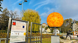 Planetenweg in Grein: Ein großer leuchtend oranger Sonnenball thront auf einem Sockel in einem gepflegten Park. Bunte Bäume, Büsche und ein aufgestellter Informationsständer rahmen die Szene ein. Am Himmel zeigen sich ein paar Wolken vor dem blauen Hintergrund.