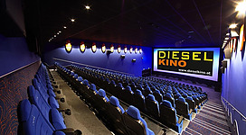 Innenansicht eines Kinosaals mit vielen blauen Sitzen. Auf der Leinwand ist das Logo ‚DIESEL KINO‘ zu sehen. Die Wände sind dunkelblau und die Decke mit kleinen Lichtern beleuchtet.