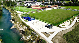 Ein Sportgelände mit einem großen Fußballfeld und mehreren anderen Sportplätzen. Neben dem Gelände fließt ein klarer Bach und es sind Bäume und grüne Flächen sichtbar.