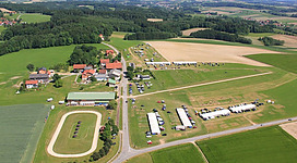 Islandpferdehof Lichtegg in Andorf: Eine ländliche Umgebung mit mehreren Gebäuden und Parkplätzen, umgeben von Feldern und Waldgebieten. Es gibt eine kreisförmige Struktur, die möglicherweise eine Rennstrecke oder Sportstätte darstellt. Insgesamt eine idyllische Landschaft mit einer Mischung aus landwirtschaftlichen Flächen und bewaldeten Gebieten.
