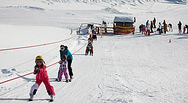 Eine lebhafte Skischule mit Kindern, die auf einer schneebedeckten Piste Skifahren lernen. Im Hintergrund sind weitere Menschen und ein Holzhaus zu sehen.