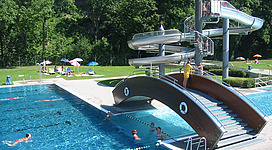 Freibad Ried im Innkreis in Ried im Innkreis: Ein großer, glänzender Wasserpark mit rutschbahnen, Schwimmbecken und Menschen, die sich dort vergnügen. Mehrere Rutschen führen in die verschiedenen Schwimmbecken, die von einer grünen Umgebung umgeben sind. Viele Besucher genießen den Spaß in den Wasserbecken.