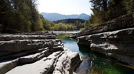 Ein ruhiger Fluss fließt zwischen felsigen Ufern mit grünen Bäumen im Hintergrund. Die Umgebung ist hell erleuchtet und vermittelt ein Gefühl von Natur und Frieden.