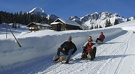 Eine winterliche Landschaft mit schneebedeckten Hügeln und Bergen im Hintergrund. Drei Personen fahren mit Schlitten einen verschneiten Hang hinunter.