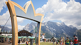 Ein großer, kreativer Holzbogen steht im Vordergrund, umgeben von vielen Menschen. Im Hintergrund sind beeindruckende Berge und ein blauer Himmel zu sehen.