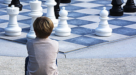 Freiluft Schach in Seewalchen am Attersee: Das Bild zeigt einen großen Schachbrett-Spielplatz mit riesigen weißen und schwarzen Spielfiguren. Ein junger Betrachter steht inmitten der Spielfiguren und betrachtet die Szene.