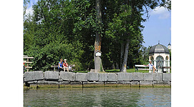 Blick vom See auf das Ufer, Personen sitzen auf einer Bank, im Hintergrund Bäume, seitlich eine kleine Seekapelle