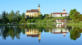 Badesee Waldhausen im Strudengau in Waldhausen im Strudengau: Eine mittelalterliche Burg mit einem hohen Turm und roten Dächern, die sich an einem See mit klarem Wasser und Bäumen am Ufer widerspiegelt. Die Landschaft ist von grünen Wäldern und Hügeln umgeben, was eine malerische und friedliche Szene ergibt.