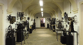 Ein langer Flur mit hohen Decken, der von alten Maschinen und Gerätschaften gesäumt ist. Die Wände sind hell und der Boden ist aus Steinen.