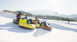 Eine Gruppe von Menschen fährt rutschend auf einer Schneebahn mit bunten Reifen. Im Hintergrund sind verschneite Berge und ein klarer Himmel zu sehen.
