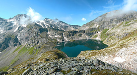 Eine beeindruckende Berglandschaft mit einem klaren See in der Mitte. Umgeben von hohen Bergen und einem blauen Himmel mit wenigen Wolken.