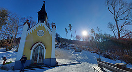 Eine kleine, gelbe Kapelle steht in einer verschneiten Landschaft. Die Sonne scheint klar am Himmel und im Hintergrund sind Bäume zu sehen.